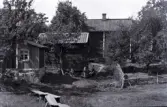 Två kvinnor skakar en matta i Stensjö bykärna. Runtom syns ett flertal byggnader och mellan dem finns inslag av ett flertal fruktträd. I bildens nedre del syns en nyslagen äng med en spång. Bakom kvinnorna är en timmrad byggnad, framför vilken det står en handdriven slipsten. På väggen hänger ett antal lieorv med monterade blad. Till höger om kvinnorna löper en trädgärdesgård.