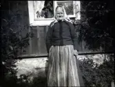 Porträtt av Titti Nilsson framför sin stuga. Titti har ett rutigt huckle och en mörk jacka och randigt förkläde. Hon bär glasögon. I bakgruden syns hennes stuga med notläkt eller fasadläkt. I fönstret syns gardiner i spets och krukväxter.