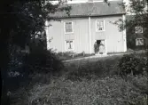 En man och Ketty Andersson framför dörren i Kettys hus i Stensjö by. Huset ör en ljus byggnad i två våningar med enkupigt tegeltak. I fönstrarna hänger gardiner. Till vänster om dörren syns en bänk. Framför huset löper en grusgång. I förgrunden på sidorna syns fruktträd och pion.