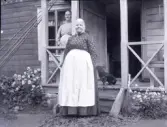 Två kvinnor framför ett hus i Stensjö by. Stående på verandan är Tilia Andersson. Till höger om henne sitter en tax. Framför verandans trappa är granris lagt och till höger om den syns en kvast av ris. På båda sidor av verandan syns perenner. På högra sidan har granris lagts mot dem och på vänstra sidan är innanför ett nät av metall. Mot verandans tak är fem metspön i bambu lutade. Till höger om den öppna dörren syns ett par träskor och på en bänk står en hink i zink.