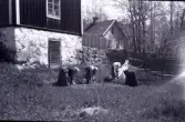 Fyra kvinnor sår lutade över såbäddar framför Undantaget i Stensjö by. Två upphöjda bara jordbäddar syns. I bakgrunden syns en samling med käppar eller kvistar. Till höger i vit hatt är möjligen Aina Stensiö. Vid knuten av Undantaget syns en kallmurad stödmur med staket ovanpå. Vid muren syns en grind i en gärdesgård av trä bunden med vidjor. I bakgrunden syns Emmas stuga och hamlade träd.