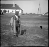 En man i utsmyckad kappa, hatt och med käpp står på en öppen plan yta. I bakgrunden syns en vit putsad byggnad och staket. Till höger syns en brunn med stång.