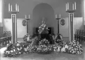 Interiörfoto från (trol. kapellet på Tegnérkyrkogården) inför en begravning. Man ser en mörk kista omgiven av höga kandelabrar och blommor.