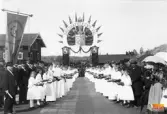 Här väntas det på Kung Oscar II den 20 augusti 1890.
Flickorna i de vita klänningarna och med vita band i håret stod ordnade i två led (84 st) och hade till uppgift att kasta blommor ur sina blomsterkorgar där kungen skulle gå fram.
Till minne av detta får varje flicka en specialtillverkad minnesmedaljong i ett etui klätt med siden och sammet i rött, som kan bäras som halssmycke eller brosch, detta är en gåva från Grevinnan von Hallwyl.
Smycket består av en präglad oval i bronsgul metall med kunglig krona och en rosenkvist i mittpartiet och med texten: Ljusne 20/8-1890 runt kanten. Ramen kring ovalen är en miniatyrkopia av Ljusnekätting, det vill säga en kätting från Ljusne kättingsmedja.
En av dessa broscher finns på Ljusne bruksmuseum.

Som 4:e flicka från vänster står greveparets dotter Irma von Hallwyl.
I den vänstra raden med flickor står som nummer sju Augusta Fager (född 1870, gift Augusta Persson, mor till Oskar Persson, Ljusne).
Mannen längst fram till vänster heter Daniel Wallin.
Vid standaret står Sjökvist som senare flyttade till Vallvik.