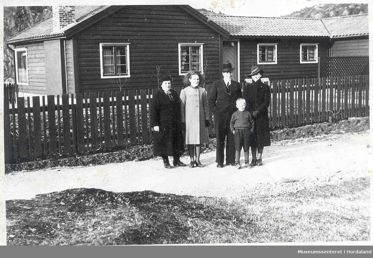 Folk i finklær, hus, tregjerde, veg, litt natur.