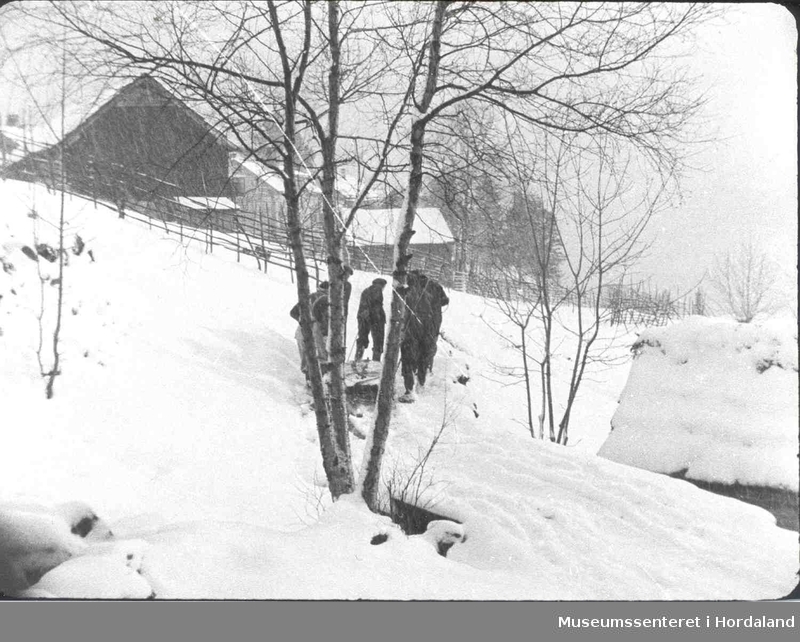 Havrå. Menn som sleper ei steinhelle på ein slede. Snødrev. Løe. Våningshus. Trehesjer.