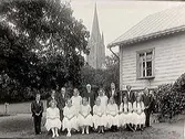 En grupp komfirmander och präst i trädgården på Årstad prästgård. I bakgrunden syns kyrkan.