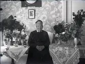 Födelsedagsporträtt av äldre kvinna. Kvinnan sitter i sitt hem omgiven av en stor mängd blombuketter i vas. Blommorna är placerade på  två bord på ömse sidor om kvinnan. Vackra dukar finns på borden. I fonden tapet med stort blommönster.