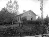 Järnvägsstation i Drängsered, Byggnaden har tegelfasad och en skylt på gaveln. En trätrappa leder ner till banområdet. I slänten står en pojke och en uniformsklädd man.