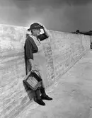 En äldre kvinna med en trälåda i handen står lutad mot en mur vid Herrviks fiskehamn, Gotland