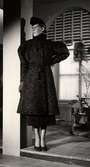 Vintermodet 1936. Kvinnlig modell poserar iförd kappa av persianpäls. Kappan har puffärmar och skärp med tofsar. I övrigt bär mannekängen vadlång rak kjol och högklackade pumps. Franska Damskrädderiet vid varuhuset Nordiska Kompaniet i Stockholm.