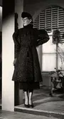 Vintermodet 1936. Kvinnlig modell poserar iförd kappa av persianpäls. Kappan har puffärmar och skärp med tofsar. I övrigt bär mannekängen vadlång rak kjol och högklackade pumps. Franska Damskrädderiet vid varuhuset Nordiska Kompaniet i Stockholm.