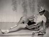 Sommarmode visas på varuhuset Nordiska Kompaniet i Stockholm 1936. En kvinnlig modell poserar, sittande i en randig strandstol av textil, iförd stranddräkt bestående av shorts, kortärmad blus och prickig keps med knytband. Hon bär nedvikta strumpor och remsandaletter. Modellen läser en tidning.