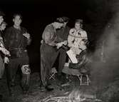 Nattorientering, män och en kvinna i mörker står runt en man som sitter på en fällstol intill en eld. Nordiska Kompaniet 1940.