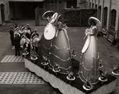 Barnens dag på Nordiska Kompaniet i september 1941. Tre mycket långa kvinnor (på styltor) i herdinneklädsel med parasoll står på en festligt dekorerad vagn förspänd med fyra hästar. Vid varje häst står en lakej. Jämför NMA.0050770.