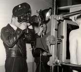 Skärmbildsfotografering på Nordiska Kompaniet 1943. En man i skinnjacka fotograferar, en annan man sköter belysningen och riktar en lampa mot en skärm, bakom vilken står en man med bar överkropp.