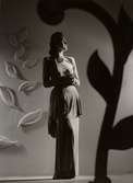 Franska avdelningen på Nordiska Kompaniet 1944. Kvinnlig modell poserar i dramatisk ljussättning iförd draperad aftonklänning och flerradig pärlcollier. På väggen bakom ett arrangemang med blad. 