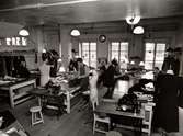 Beklädnadsateljén vid varuhuset Nordiska Kompaniet i Stockholm 1947. Män och kvinnor arbetar med sömnad vid långa arbetsbord i skrädderiet. De syr för hand, men i rummet står också två symaskiner av fabrikatet Singer. Där står också några provdockor, så kallade mannekänger.