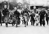 Barn, små kullor och masar, i folkdräkt springer på väg mot fotograf. Barnen kallar till Valborgsmässa 30 april 1836 i Floda genom att ringa i koskällor.