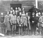 Gruppbild föreställande män boende i Ilomants, Karelen, Finland. Männen som troligen är tingsfolk står framför en stuga av liggtimmer.