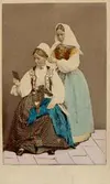 Dräktdockor med huvuden skulpterade av C A Söderman, visade på världsutställningen i Paris 1867. Två kvinnor i folkdräkter från Blekinge, varav en hjälper den andra med huvudbonaden. Handkolorerad version.