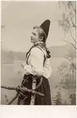 Porträtt av ung kvinna iklädd folkdräkt från Dalarna som står lutad mot ett staket. Handkolorerat fotografi.