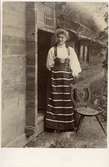En kvinna poserar i folkdräkt från Dalarna utanför en stuga. Krönt allmogestol.