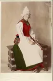 Porträtt av kvinna klädd i helgdagsdräkt för gift kvinna från Ore socken i östra Dalarna. Handkolorerat foto.