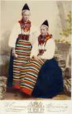 Två unga kvinnor poserar i folkdräkter från Rättvik i Dalarna. den ena kvinnan är möjligen Vass Anna. Kolorerat fotografi.
