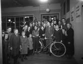 Gruppbild, barn, mest pojkar omkring en cykel. Tävling på Nordiska kompaniet för skolungdom. 