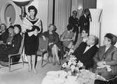 Pälsuppvisning på Nordiska Kompaniet, Franska avdelningen. En kvinnlig modell visar en stola av ljus päls. Vid små bord sitter kvinnor och och män och ser på. 2 november 1959.