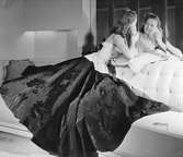 Aftonklänning från Franska avdelningen på varuhuset Nordiska Kompaniet. En kvinnlig modell sitter i en soffa, vänd mot en spegel bakom soffryggen. I handen håller hon en pudervippa som hon för mot ansiktet. Klänningen är ärmlös och den vida blommönstrade kjolen ligger draperad.