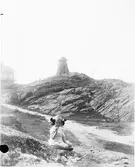 Flicka sitter på berg iklädd klänning och rosetter i håret. Kvarn i bakgrunden. Fiskebäckskil, Bohuslän.