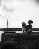 Två flickor på klipphäll, i bakgrunden segelbåt. Troligen i Fiskebäckskil, Bohuslän.