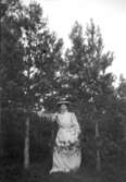 Helporträtt av en kvinna i vit klänning och hatt bland tallar.