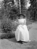 En gammal kvinna i vitt förkläde och rutig blus sitter framför en blomrabatt.