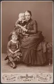 Kabinettsporträtt av kvinna (Henrietta Maria Carlson f. Tempelman f. 7/6 1860 d. 23/6 1950) gift med Fredrik Carlson (1854-1927) med två barn (kapten Arne Carlson f. 20/10 1881 d. 17/9 1921 och musikdirektör Sigrid Carlson f. 9/12 1883 d. 9/7 1935). Kvinnan möjligen sorgklädd bärande sorgsmycken i form av grovt halsband och armband.
Grupporträtt.