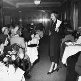 Konfektionsvisning i lunchrummet på Nordiska Kompaniet.
Modell passerar publiken, klädd i svart kappa, svart hatt, dubbelt pärlhalsband och svarta pumps. Modellen håller en skylt som visar 295 SEK. Publiken sitter vid uppdukade bord med vita dukar, NK-servis och blommor i vas. De flesta kvinnorna i publiken bär hatt.