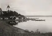 Rättviks kyrka i Dalarna. Kyrkbåtar på Siljans vatten. Vid stranden framför kyrkstallarna står högtidsklädda människor.
