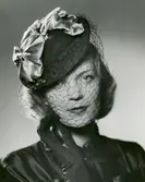 Porträtt av kvinna klädd i mörk sidenblus, mörka handskar, samt  hatt med svart flor och ljusa rosetter i siden.