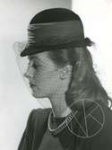 Porträtt av kvinna i hatt med brett hattband och flor, samt halsband.