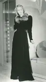 Mannekäng, Margit Langlet, i lång kappa av sammet med pälskrage, spegeldörrar och lampa i bakgrunden. NK:s Franska damskrädderi 1939.