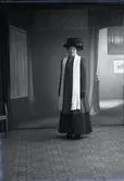 Kvinnoporträtt, kvinna stående i helfigur iförd ytterkläder, hatt och halsduk. Ateljébild. Beställare: Anna Svensson.
