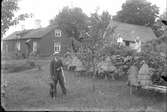En man med jaktgevär och hund, en kvinna och en flicka i en trädgård framför ett bostadshus.