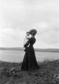 En kvinna med ett barn i famnen står på stranden och tittar mot Byfjorden i bakgrunden