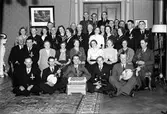 Kvinnor och män poserar tillsammans, några med musikinstrument i främre raden. Vissa män är dekorerade med ett kors med texten (troligen) ISHF 1908-1938.