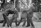 Fem män varav fyra engagerade i låtsasbrottning respektive låtsasboxning, i bakgrunden bänkar, en gräsbevuxen vall samt träd. Vid Lungkliniken, Eksjö.
Fotografens anmärkning: 