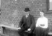Gustav Anderssons far och syster sitter på en bänk utmed en tegelvägg.