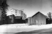 Enköping, uthus på stadsägorna 231 och 225 - Kölnbacksgatan  1 A  och 1 B, från Fjärdhundragatan, mot nordväst. I bakgrunden punkthusen i kvarteret  Påfågeln. Januari 1957.