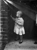 En liten flicka i klänning och kofta står och känner på en tegelvägg. Bakom henne syns en dörr.